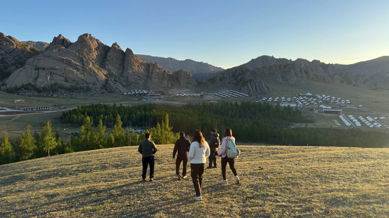 Travellers walking across a grassy hillside at sunset below granite peaks in central Mongolia, late May 2026